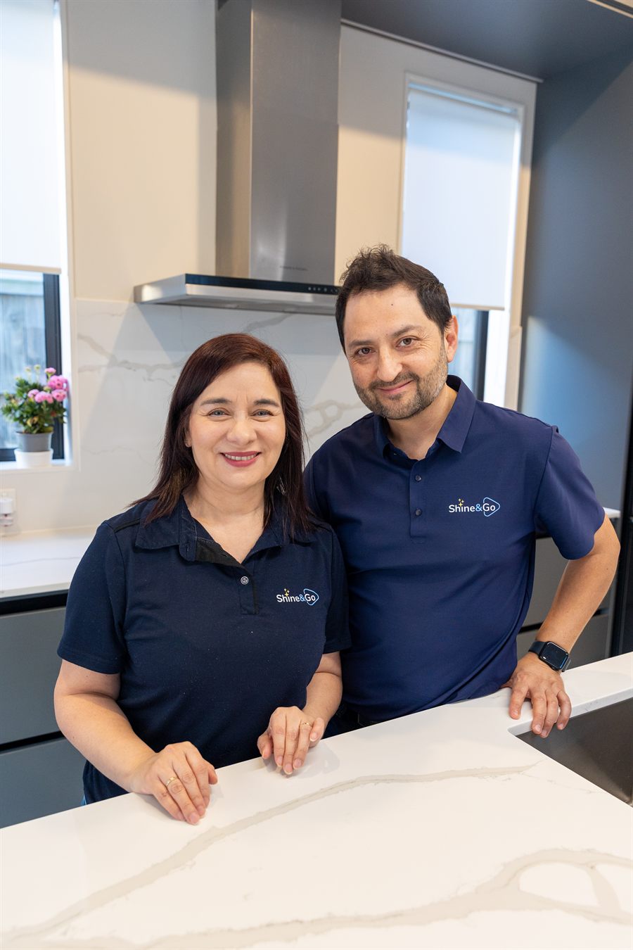 Elba and Leo, founders of Shine&Go NZ, in a kitchen wearing branded Shine&Go polo shirts